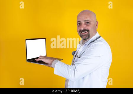 cheerful middle aged European male doctor holding, typing laptop with white blank screen for mock up. Looking camera in workwear using pc notebook. Stock Photo