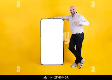 Mobile advertisement concept. Cheerful middle aged caucasian male leaning and pointing big modern mobile phone with empty screen for mockup. Stock Photo