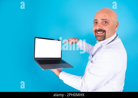 Portrait of middle aged doctor showing laptop. Isolated blue background. General practitioner holding notebook with empty screen pointing display. Stock Photo