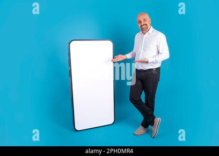 Caucasian bald middle aged man recommending new mobile app advertisement. Full body shot male with big smartphone empty screen, mockup. Stock Photo