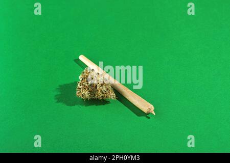 A king size joint lies next to dry marijuana buds on a green background.  Lots of empty space Stock Photo