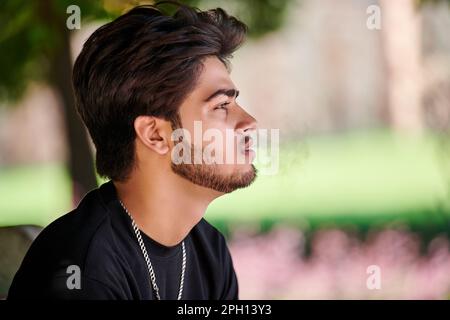 Attractive indian man smoker exhales cigarette smoke portrait in black t shirt and silver neck chain in public park, hindu male smoking close up portr Stock Photo