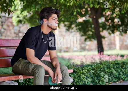 Attractive indian man smoker exhales cigarette smoke portrait in black t shirt and silver neck chain sitting on bench in public park, hindu male smoki Stock Photo