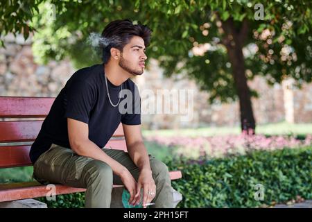 Attractive indian man smoker exhales cigarette smoke portrait in black t shirt and silver neck chain sitting on bench in public park, hindu male smoki Stock Photo