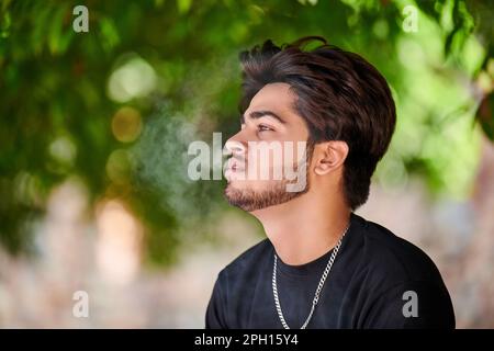 Attractive indian man smoker exhales cigarette smoke portrait in black t shirt and silver neck chain in public park, hindu male smoking close up portr Stock Photo