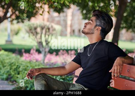 Attractive indian man smoker exhales cigarette smoke portrait in black t shirt and silver neck chain sitting on bench in public park, hindu male smoki Stock Photo