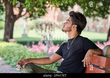 Attractive indian man smoker exhales cigarette smoke portrait in black t shirt and silver neck chain sitting on bench in public park, hindu male smoki Stock Photo