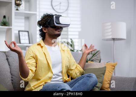 Young Hindu man in yellow shirt alone at home sitting on sofa and meditating in lotus position, man using vr glasses for virtual meditation and travel. Stock Photo