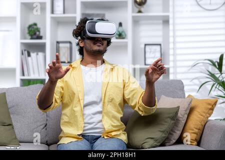 Latin American man sitting on couch at home using virtual reality glasses, man viewing educational material remotely. Stock Photo