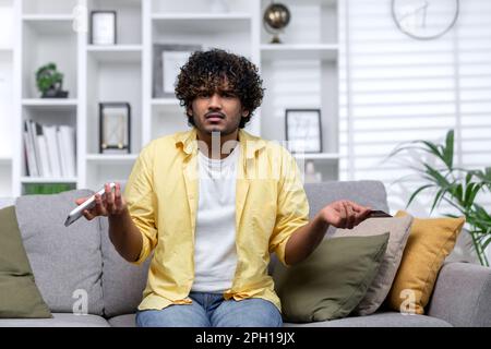 Portrait of cheated online shopper at home, man upset looking at camera, hispanic holding phone and bank credit card, missing stolen money on account, fraudulent actions. Stock Photo
