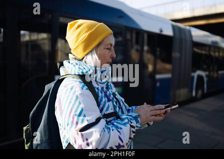 Old woman traveler with luggage watches the train schedule and buys train tickets using electronic application by her smartphone while waiting on the Stock Photo