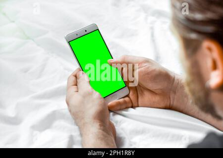 Young Bearded Man Using Green Chroma Key Screen Phone, Relaxing in a Bed. Guy Uses Green Mock-up Screen Smartphone. Rear View. Man lying on white blan Stock Photo