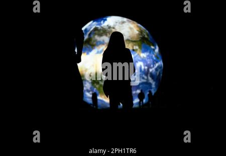 Silhouette of young woman standing in front of large inflatable model of planet Earth glowing at night, view form behind Stock Photo