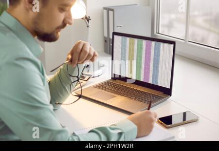 Side view of young business man working on a laptop with tables at office and writing. Stock Photo