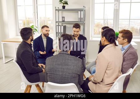 Smiling young colleagues sitting in circle are having fun conversation at business training with trainer. Stock Photo