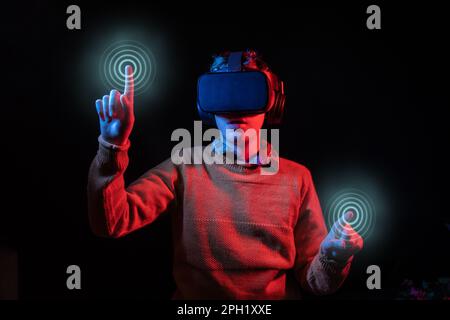 Man wearing a VR headset, pressing virtual points with both index fingers using hand tracking; studio shot with blue and red lights on black backgroun Stock Photo