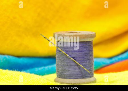 Spool of purple cotton thread and sewing needle sitting on bright colored clothing. Textile tailoring concept with focus on foreground. Stock Photo