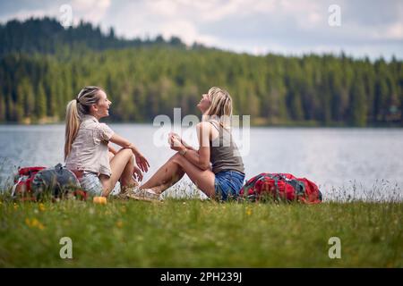 Young female friends are sitting on the ground beside the lake while hiking the hills on a beautiful day. Trip, nature, hiking Stock Photo