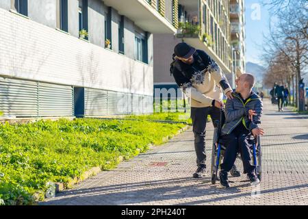 A young man in a wheelchair being assisted by a companion while making his way along the sidewalk Stock Photo