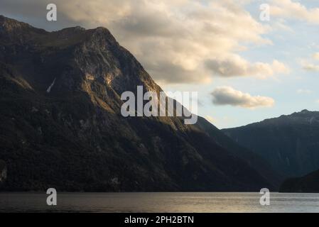 Evening light illuminates the towering mountains and cliffs that line the Doubtful Sound in New Zealand. Stock Photo