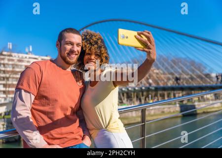 A young couple wearing vibrant outfits taking selfie outdoors in an urban area on a sunny day Stock Photo