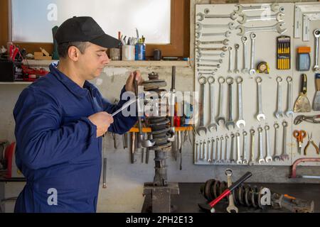 Image of a mechanic in overalls in his workshop while he disassembles a car shock absorber with a wrench and extractors. Stock Photo
