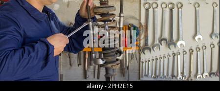 Image of a mechanic in overalls in his workshop while he disassembles a car shock absorber with a wrench and extractors. Horizontal banner Stock Photo