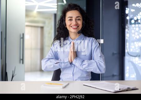 A young Muslim woman sits in the office at a desk in front of a camera in a headset. She put her hands together, thanks, says hello, communicates. Online interview, work, study. Stock Photo