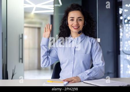 A young Muslim woman sits in the office at a desk in front of a camera in a headset. Greets, waves at the camera, communicates. Online interview, work, study, podcast recording, blog. Stock Photo