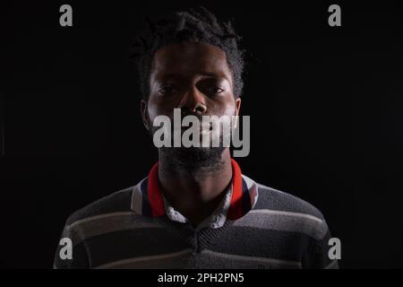Frontal headshot of a serious young African man with short braided hair, wearing a striped sweater, looking at the camera against a black background Stock Photo