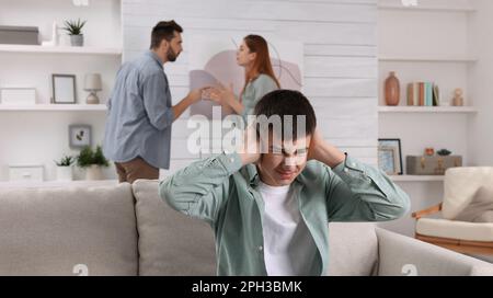 Unhappy teenage boy covering ears while his parents arguing on background. Problems at home Stock Photo