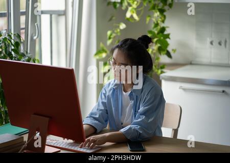 Focused Asian woman IT developer wearing glasses writing code on computer while working from home Stock Photo