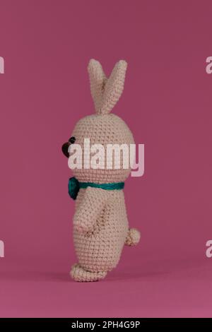 Symbol of Catholic Easter. Grey cute Easter bunny with green bow tie crocheted, handmade art. Side view. Amigurumi rabbit doll on pink background. Sof Stock Photo