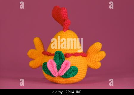 Orange cockerel with bright red comb on its head crocheted, handmade art. Amigurumi doll chicken with multicolored tail on pink background. Soft DIY t Stock Photo