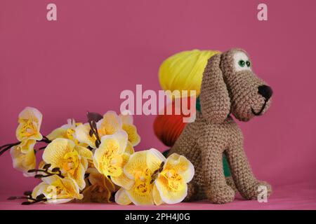 Amigurumi puppy doll on pink background next to yellow orchid flowers and multicolored wool balls. A soft DIY toy made of cotton. One brown dog Stock Photo