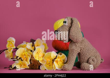 Amigurumi puppy doll on pink background next to yellow orchid flowers and multicolored wool balls. A soft DIY toy made of cotton. One brown dog Stock Photo