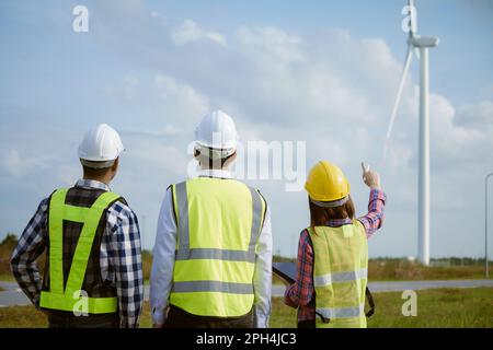 Back view of three engineers discussing and checking turbines on wind turbine farm. Renewable energy technology and sustainability. Stock Photo