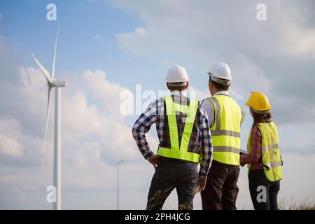 Back view of three engineers discussing and checking turbines on wind turbine farm. Renewable energy technology and sustainability. Stock Photo