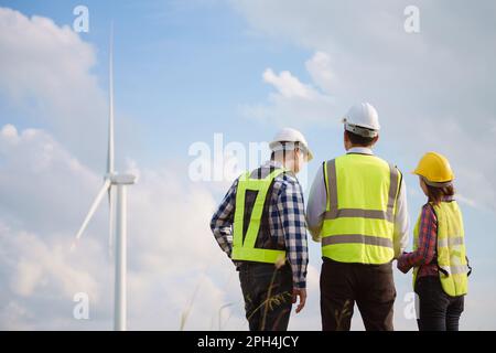 Back view of three engineers discussing and checking turbines on wind turbine farm. Renewable energy technology and sustainability. Stock Photo