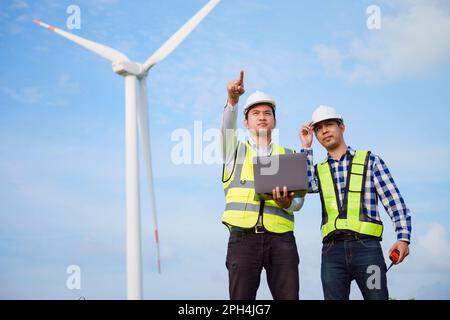Two asian engineers discussing and checking turbines on wind turbine farm. Renewable energy technology and sustainability. Stock Photo