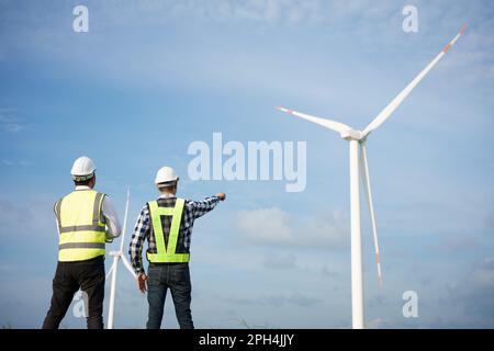 Two asian engineers discussing and checking turbines on wind turbine farm. Renewable energy technology and sustainability. Stock Photo