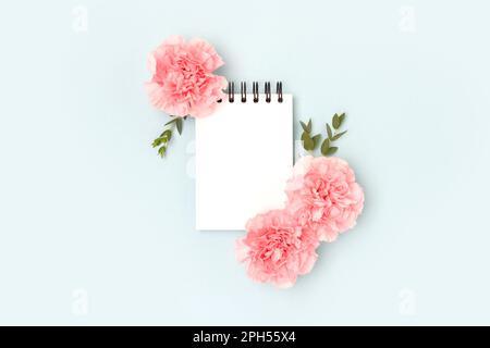 Blank notepad mockup, pink carnation flowers and green eucalyptus branches on a blue background. Place for text. Stock Photo