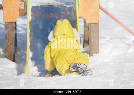 Toddler baby boy rides down a slide playing on a winter playground. A child in a yellow jumpsuit on a children's slide in the snow. Kid age one year e Stock Photo
