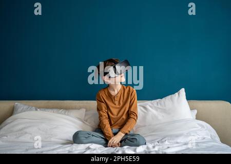 Interested schoolboy sit on bed look in 3D virtual reality glasses for smartphone immersive game Stock Photo