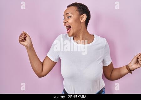 Beautiful african american woman standing over pink background dancing happy and cheerful, smiling moving casual and confident listening to music Stock Photo