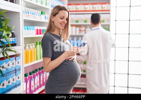 Man and woman pharmacist and pregnant client using smartphone speaking at pharmacy Stock Photo