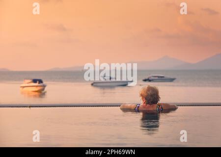 Active senior woman in swimming pool. Rear view of woman enjoying relax and watching sea in sunset. Stock Photo