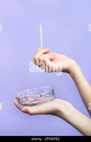 Female hands with natural nails hold a joint with medical marijuana and an empty glass ashtray on a light purple background.  copy space Stock Photo