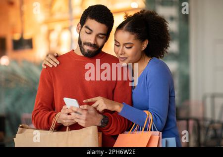 Multiethnic Spouses Shopping Using Cellphone Pointing Finger Standing At Mall Stock Photo