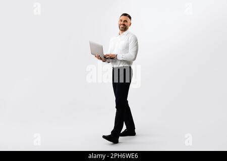 Full length shot of businessman with laptop walking over light grey studio background, male entrepreneur using computer Stock Photo